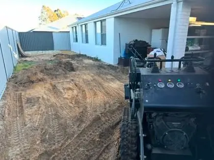 Mini loader working down a narrow side yard beside the house