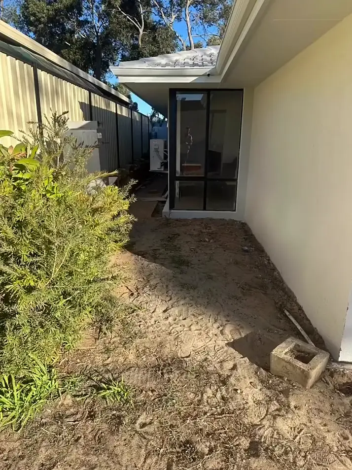 Side path along a house, bare dirt and garden overgrowth before paving